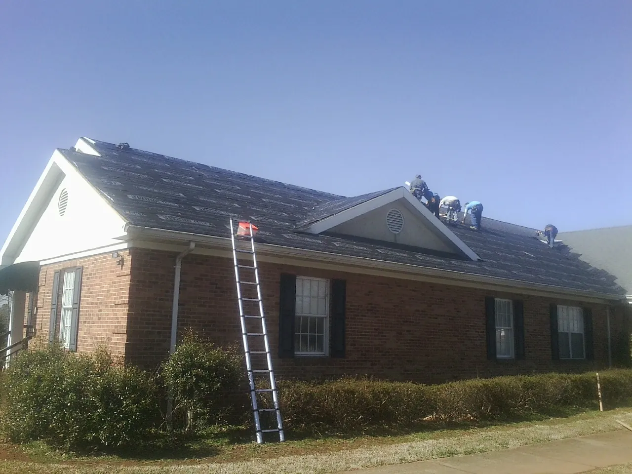 Crew tearing shingles off a brick ranch roof with a ladder set — Emergency Roof Repair work by our crew in Glen Saint Mary