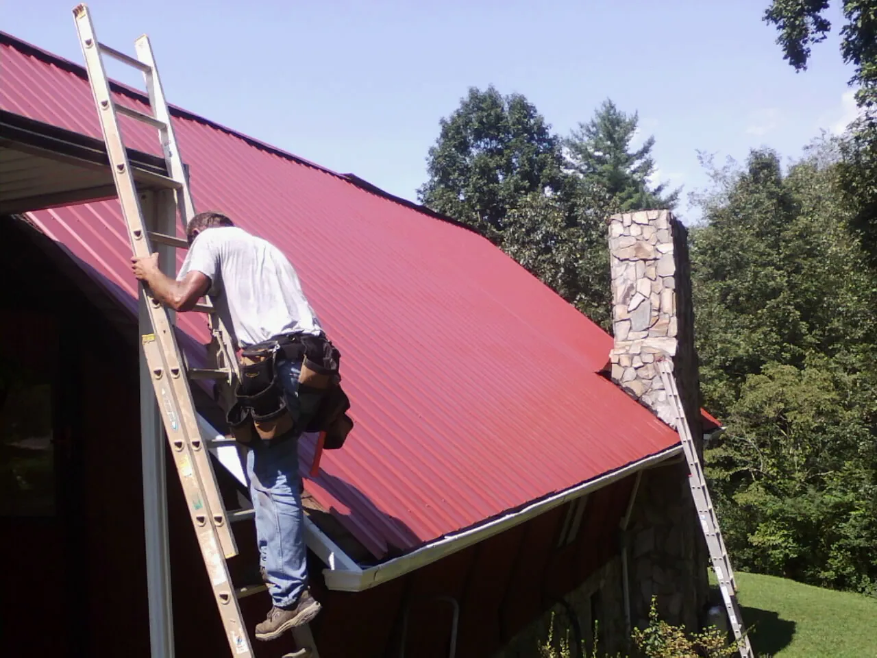 Roofer on a ladder beside a red metal roof and stone chimney — Soffit & Fascia Repair by Roofing Brothers in Glen Saint Mary
